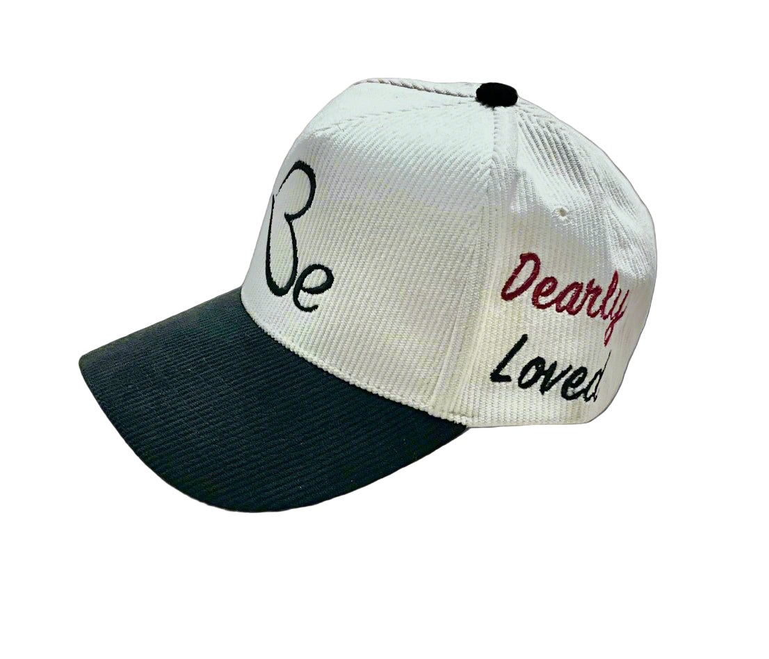 Cream and black cap with the text 'Be' and 'Dearly Loved' embroidered on it, displayed in side-view.